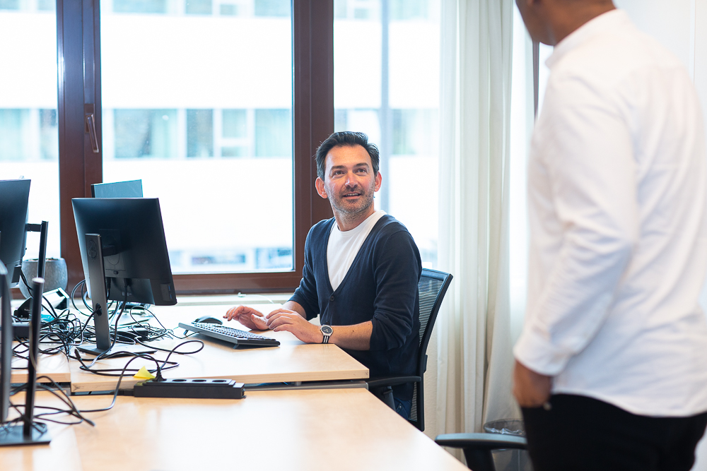 twee collega's zijn in gesprek. Een man zit aan een bureau de andere man staat